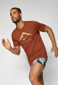 Herre rustfarget Nike Trail t-skjorte med et gull logo, kombinert med grå mønstredeshorts. Designet for aktiv bevegelse og komfort.