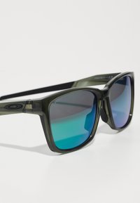 Gafas de sol verdes translúcidas con montura cuadrada, lentes en degradado de azul a púrpura y acentos laterales; material plástico liviano.
