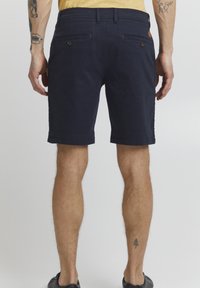 Marineblaue Shorts aus Baumwolle mit glatter Textur, ausgestattet mit zwei Gesäßtaschen und einem umgeschlagenen Saum, getragen von einer Person von hinten.