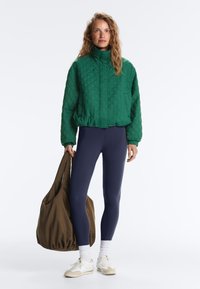 Femme portant une veste matelassée verte et des leggings bleu marine, tenant un grand sac marron, debout devant un fond blanc uni.