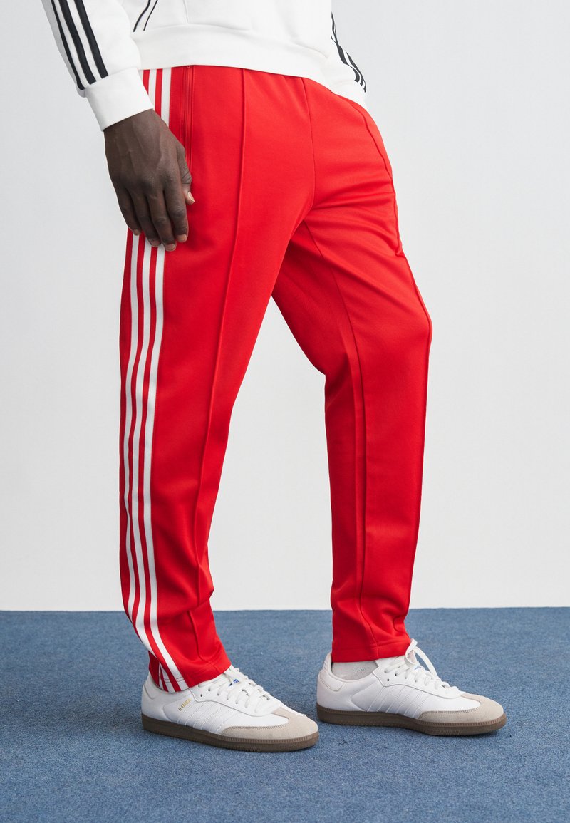 Červené sportovní kalhoty s bílými bočními pruhy a zúženým střihem. V kombinaci s bílými teniskami outfit zdůrazňuje sportovní design.