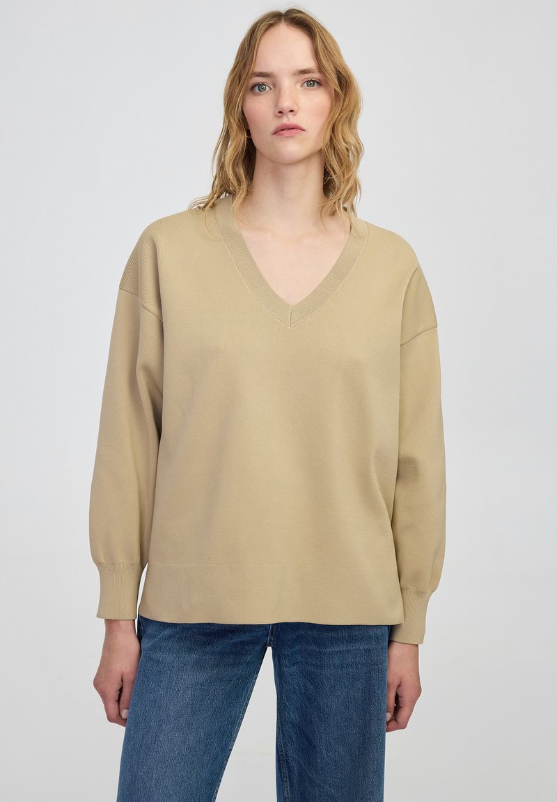 Beżowa bluza z dekoltem w serek z prążkowanego materiału i długimi rękawami, w połączeniu z niebieskimi dżinsami o prostym kroju. Prosty design z luźnym dopasowaniem.