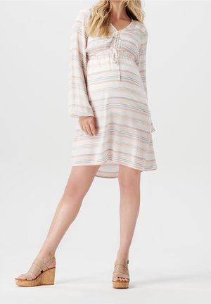 Gravid kvinde iført en knælang, pastelfarvet stribet kjole med lange ærmer og beige kile-sandal med korkhæl.