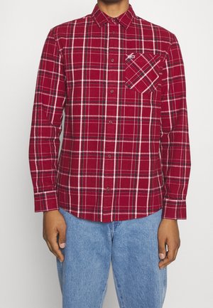 Hombre que lleva una camisa de cuadros roja de manga larga con un bolsillo en el pecho y jeans azul claro, de pie contra un fondo simple.