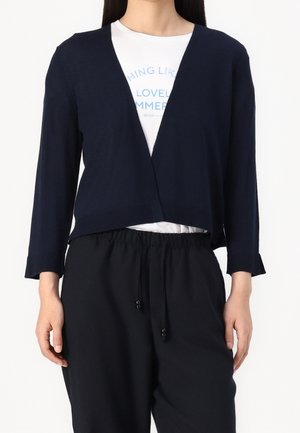 Kvinde iført en marineblå cropped cardigan over en hvid t-shirt med grafik og sorte bukser med snøre i taljen, stående mod en ensfarvet hvid baggrund.
