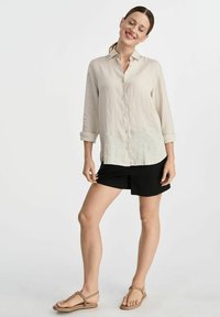 Jasno beżowa lniana koszula z zapięciem na guziki, z podwiniętymi rękawami, w połączeniu z czarnymi szortami i brązowymi sandałami. Prosty i swobodny design.