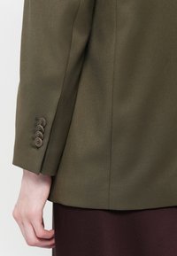 Olivově zelený blazer s hladkou texturou, s manžetou na knoflík se čtyřmi hnědými knoflíky a přiléhavým střihem.