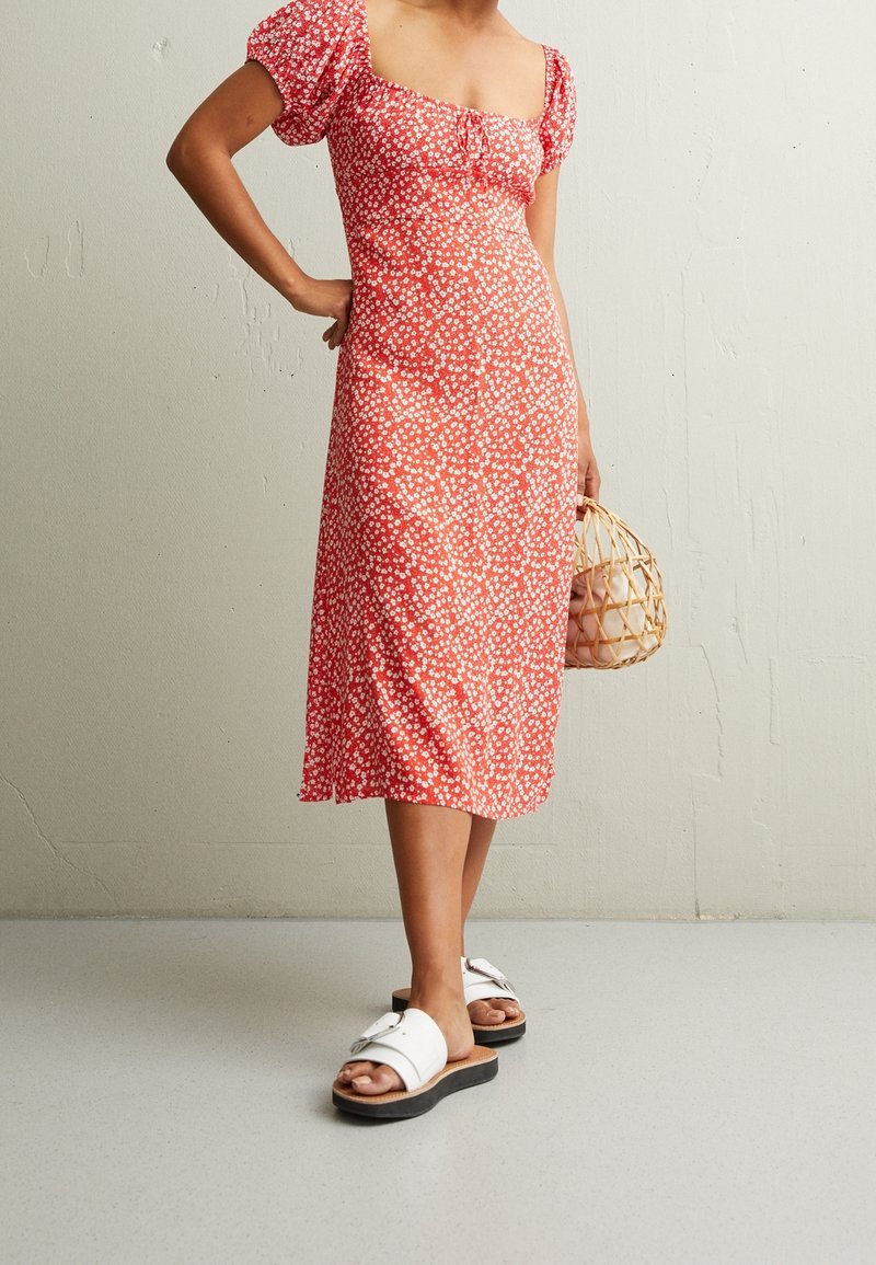 Robe midi rouge à fleurs avec manches bouffantes et décolleté carré. Paire de sandales blanches avec une semelle épaisse en bois et un sac tissé.