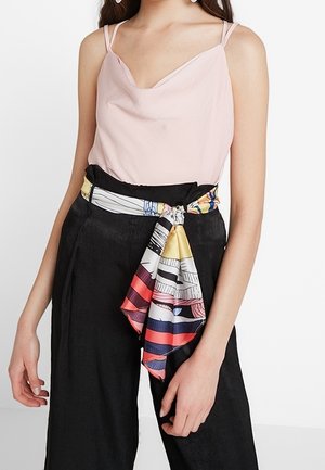 Mujer con una camiseta sin mangas de color rosa pálido metida en pantalones negros de cintura alta con un cinturón de pañuelo colorido y estampado atado en la cintura.