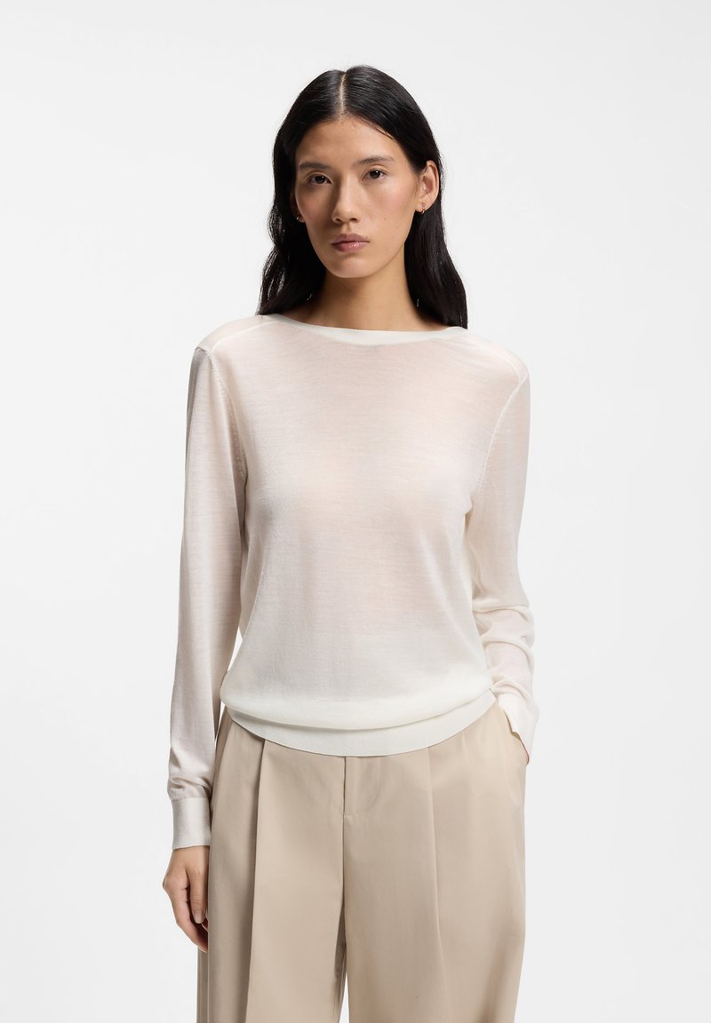 Femme aux longs cheveux foncés portant un haut blanc transparent à manches longues et un pantalon beige taille haute, debout devant un fond uni.