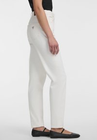 Jeans de mezclilla blanca con un corte recto, bolsillos cosidos y un parche de logo triangular en la parte trasera. Llevados con bailarinas negras, con correas en el tobillo.