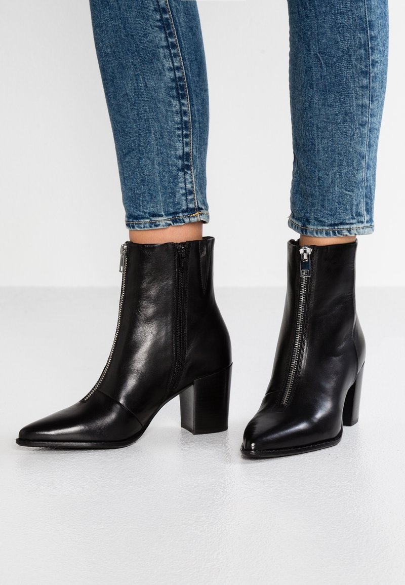 Botas de cuero negras hasta el tobillo con cremalleras plateadas frontales y tacones cuadrados, usadas con jeans azules remangados sobre un suelo blanco.