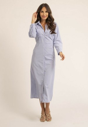 Femme aux longs cheveux bruns portant une robe midi à rayures bleues claires boutonnée et des talons beige à lanières, posant avec un léger sourire.