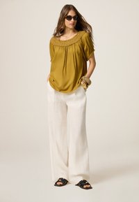 Olijfgroene top met gerimpelde halslijn, gecombineerd met losvallende witte linnen broek en zwarte sandalen. Accessoires omvatten gouden armbanden.