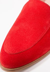 Gros plan d’une chaussure loafer en daim rouge avec des coutures visibles et une semelle marron clair.