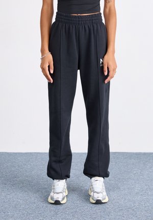 Personne portant un pantalon de survêtement noir à taille élastique, des baskets blanches, un top court noir, avec un tatouage sur le bras gauche et des bracelets au poignet droit.