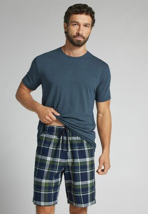 Mann mit Bart, der ein blaues T-Shirt und grün-navy karierte Shorts trägt und sein Shirt leicht anhebt vor einem einfarbigen grauen Hintergrund.