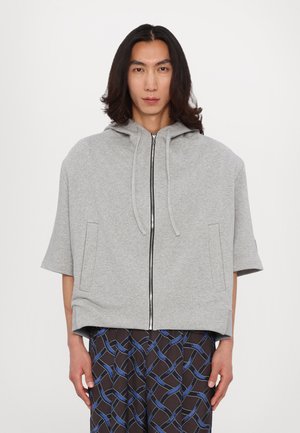 Grauer Kapuzenpullover mit kurzen Ärmeln, ausgestattet mit einem Frontreißverschluss, zwei Seitentaschen und einer lockeren Passform. Kombiniert mit gemusterten schwarzen Hosen.