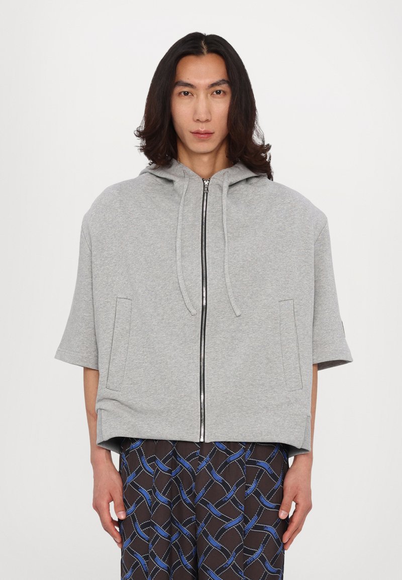 Gray hooded sweatshirt with short sleeves, featuring a front zipper, two side pockets, and a relaxed fit. Paired with patterned black pants.