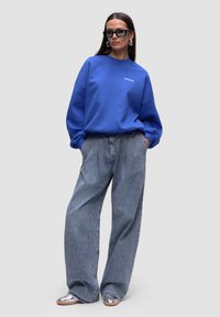 Felpa blu oversize con collo a giro, abbinata a jeans a gamba larga blu chiaro e scarpe appiattite argentate. Gli accessori includono occhiali da sole scuri.