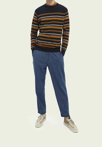 Man som bär en brun, svart och vit randig tröja, blå denimbyxor och beige sneakers, står med händerna i fickorna.