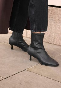 Black leather ankle boots with a pointed toe and stiletto heel. Smooth texture, minimal stitching, and slim silhouette. Worn with dark denim.