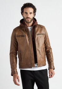 Ruskea nahkatakki, jossa on kulutettu olkatyynytyyli, vetoketjulliset taskut ja tyköistuva leikkaus. Yhdistetty ruskean hupparin kanssa.