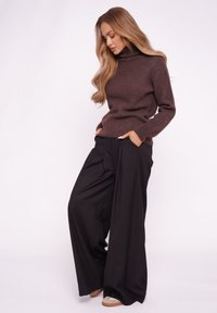 Brązowy golf z miękkiego materiału, zestawiony z szerokimi czarnymi spodniami. Model stoi z rękami w kieszeniach, prezentując strój.