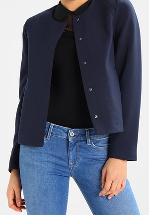 Woman wearing a navy blue buttoned jacket over a black top with a collar, paired with high-waisted blue jeans, standing with one hand relaxed.