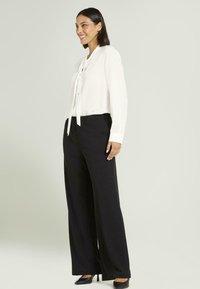 Blusa blanca con cuello atado, combinada con pantalones negros de pierna ancha de talle alto. Textura suave y ajuste entallado, complementados con tacones negros.