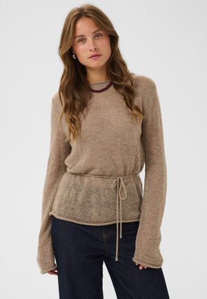 Kvinde med langt bølget hår iført en beige sweater med bånd i taljen og mørkeblå jeans mod en ensfarvet lys baggrund.