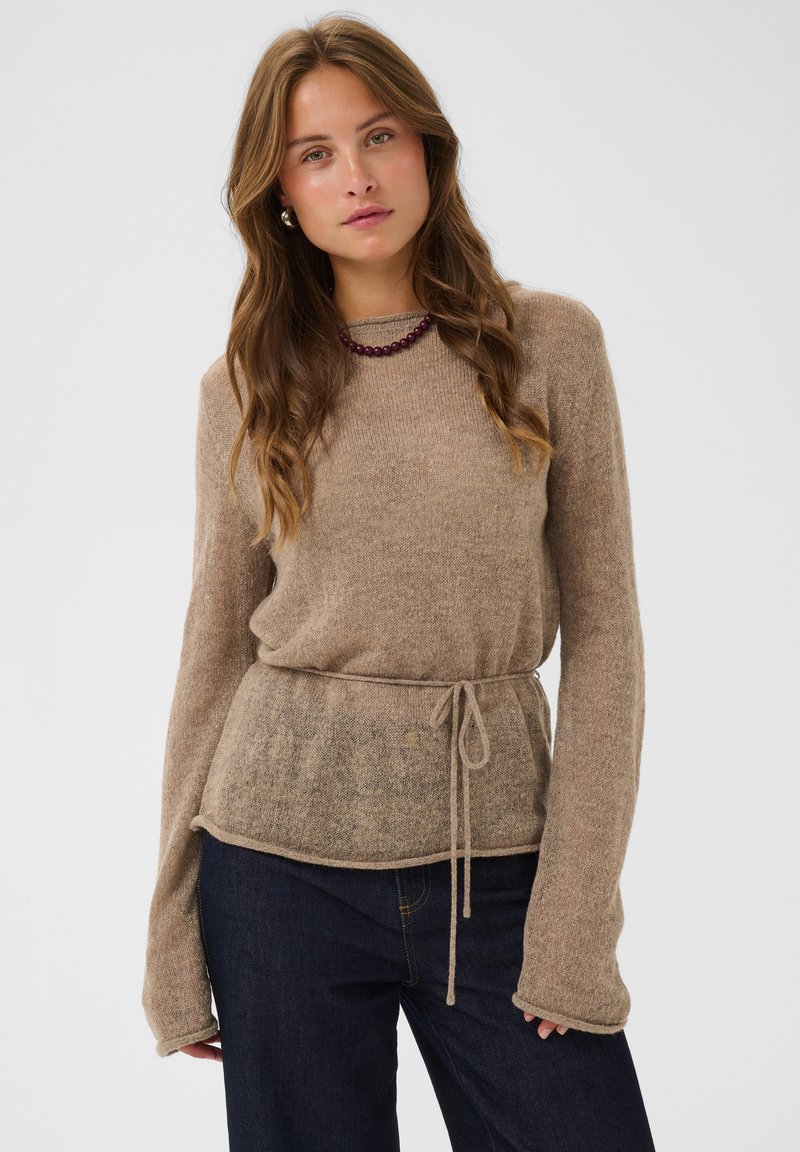 Femme aux longs cheveux ondulés portant un pull beige avec une ceinture à la taille et un jean bleu foncé sur un fond uni clair.