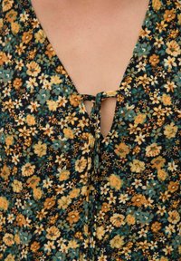Robe fleuri avec un fond noir ornée de petites fleurs jaunes, oranges et vertes. Elle a un décolleté en V et un détail accentué noué.