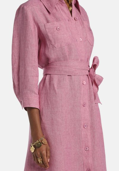 Frau trägt ein langärmliges, rosa Kleid mit Knopfleiste, Brusttaschen und Stoffgürtel, accessorisiert mit einem goldenen Armband und Ringen.