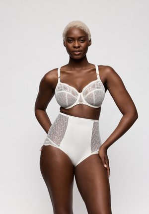 Vrouw met kort blond haar die een witte kanten beha en een slip met hoge taille draagt, staand met één hand op haar heup tegen een grijze achtergrond.