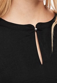 Svart blus med rund halsringning och en liten nyckelhålsöppning som hålls fast av en glänsande silverknapp. Mjuk tyg med en slät textur.