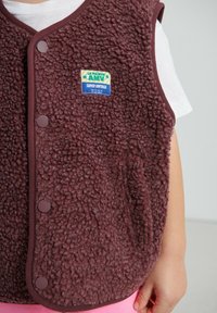 Bruine fleecevest met zachte textuur, voorzien van een blauw-groei label, drukkers en een contrasterende bies. Gedragen over een wit shirt.