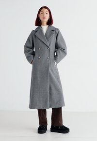 Double-breasted gray wool coat with wide lapels and two large buttons, paired with brown pants and dark shoes. Smooth texture and tailored fit.