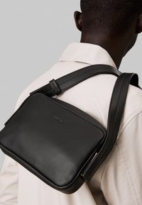 Black leather crossbody bag with a smooth texture, rectangular shape, adjustable strap, and a subtle logo on the front.