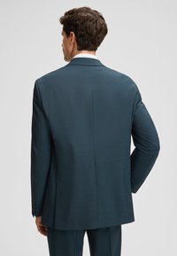Man met kort krullend haar in een donker teal colbert en bijpassende broek, met de rug naar de camera tegen een lichte, effen achtergrond.