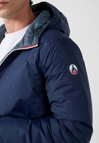 Hombre con chaqueta acolchada azul marino con capucha y parche con el logo de JOTT en la manga izquierda, camisa blanca debajo, fondo claro liso.