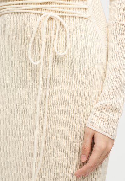 Gros plan sur une main reposant sur une robe beige en maille avec un nœud à la taille, montrant le tissu texturé et le détail de la manche longue.