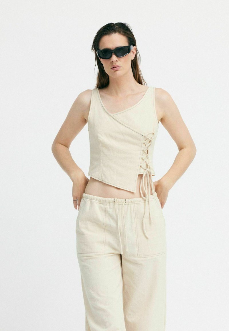 Beige Cropped-Top mit V-Ausschnitt und Schnürdetails an der Seite, kombiniert mit passenden lockeren Hosen. Glatte Stoffstruktur.
