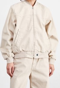 Veste bomber beige en coton avec fermeture éclair devant, poignets et ourlet côtelés. Comprend des poches latérales et un petit patch logo à la taille.