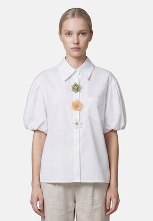 Femme portant un chemisier blanc à manches courtes bouffantes avec trois épingles décoratives en forme de fleurs au centre devant.