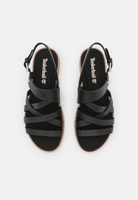 Mustat sandaalit, joissa on ristikkäiset synteettiset hihnat, pehmeä musta pohja ja vaaleanruskea kumipohja. Säädettävä nilkkahihna metalliosilla.