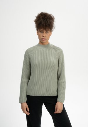 Lichtgroene ribgebreide trui met een hoge hals en lange mouwen, gemaakt van een textuur gebreide stof, gecombineerd met zwarte broeken.