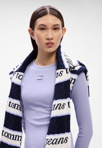 Lila gerippter Langarmtop mit einem gestreiften schwarz-weißen Schal, der ein strukturiertes Design aufweist und den Text 'TOMMY JEANS' in kontrastierenden Farben trägt.