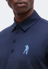 Gros plan d'un homme portant un polo bleu marine boutonné avec un petit logo silhouette bleu clair représentant une personne debout faisant le salut sur la poitrine.