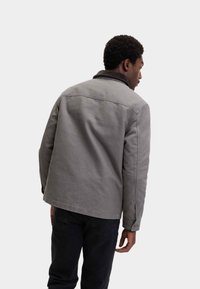 Grå denimjakke med krave, synlige sømmer og en afslappet pasform. Båret med sorte bukser. Vist bagfra.